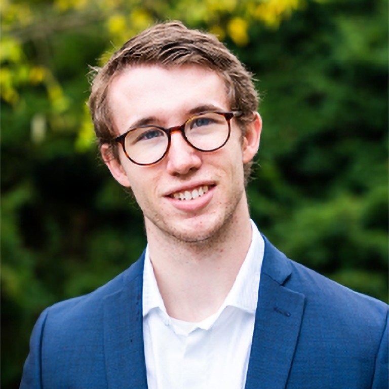 Headshot of a man smiling. He is wearing round framed glasses, a blue suit jacket and white collared shirt underneath.