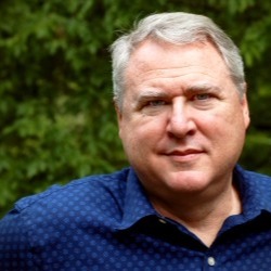 Headshot of older man wearing a dark blue shirt. He stands in front of a backdrop of green leaves