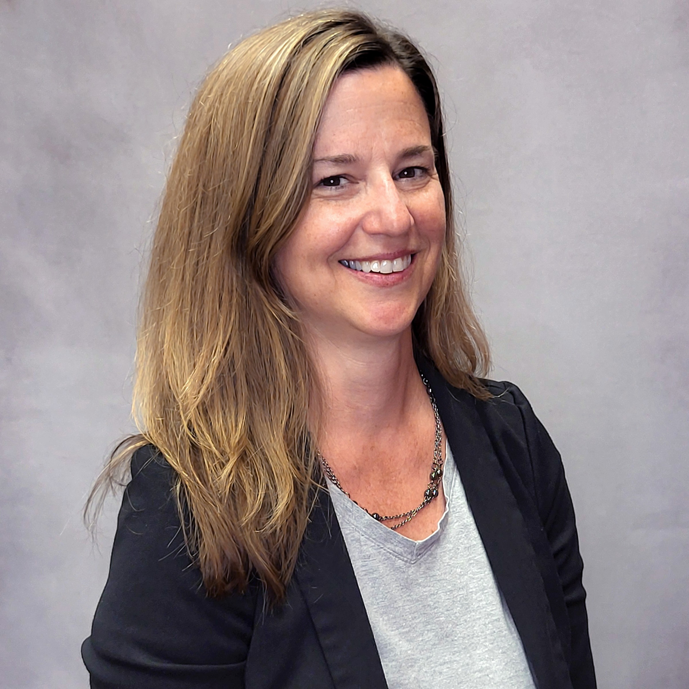 Headshot of a woman smiling in front of a gray backdrop. She is wearing a gray shirt under a black cardigan.