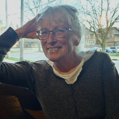 Photo of woman smiling in front of a window showing a tree and buildings outside behind her.