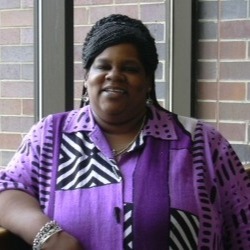 Photo of woman smiling. She is wearing a purple and black and white patterned shirt and is standing in front of a brick wall.