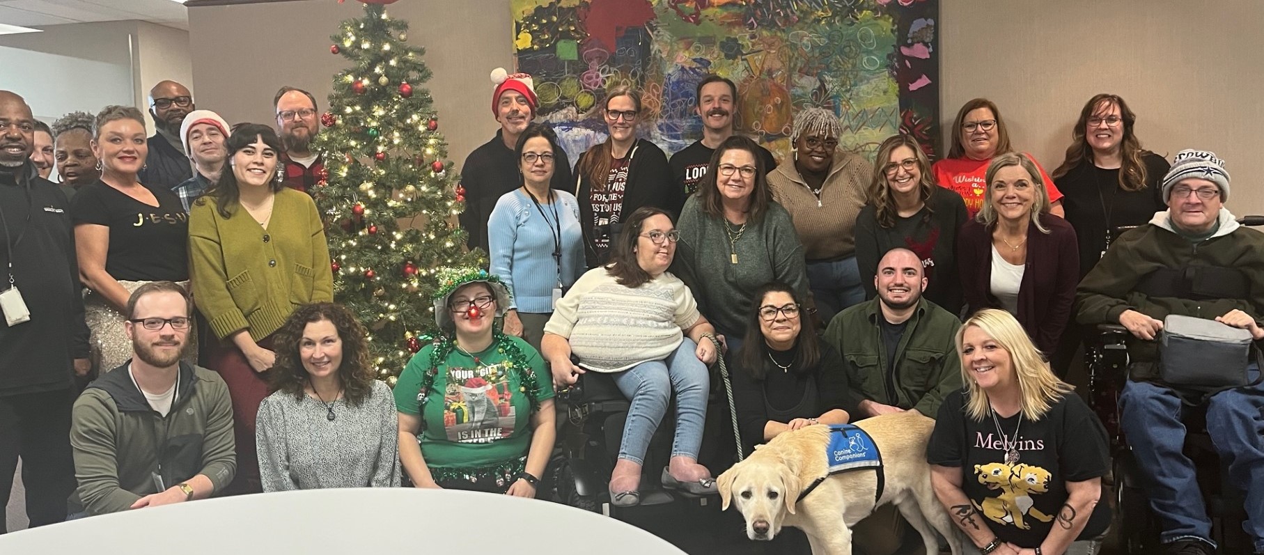 Group of 26 employees and one service dog, some dressed festively, stand, kneel, and sit together with a Christmas tree among them and a colorful painting on the wall behind them.