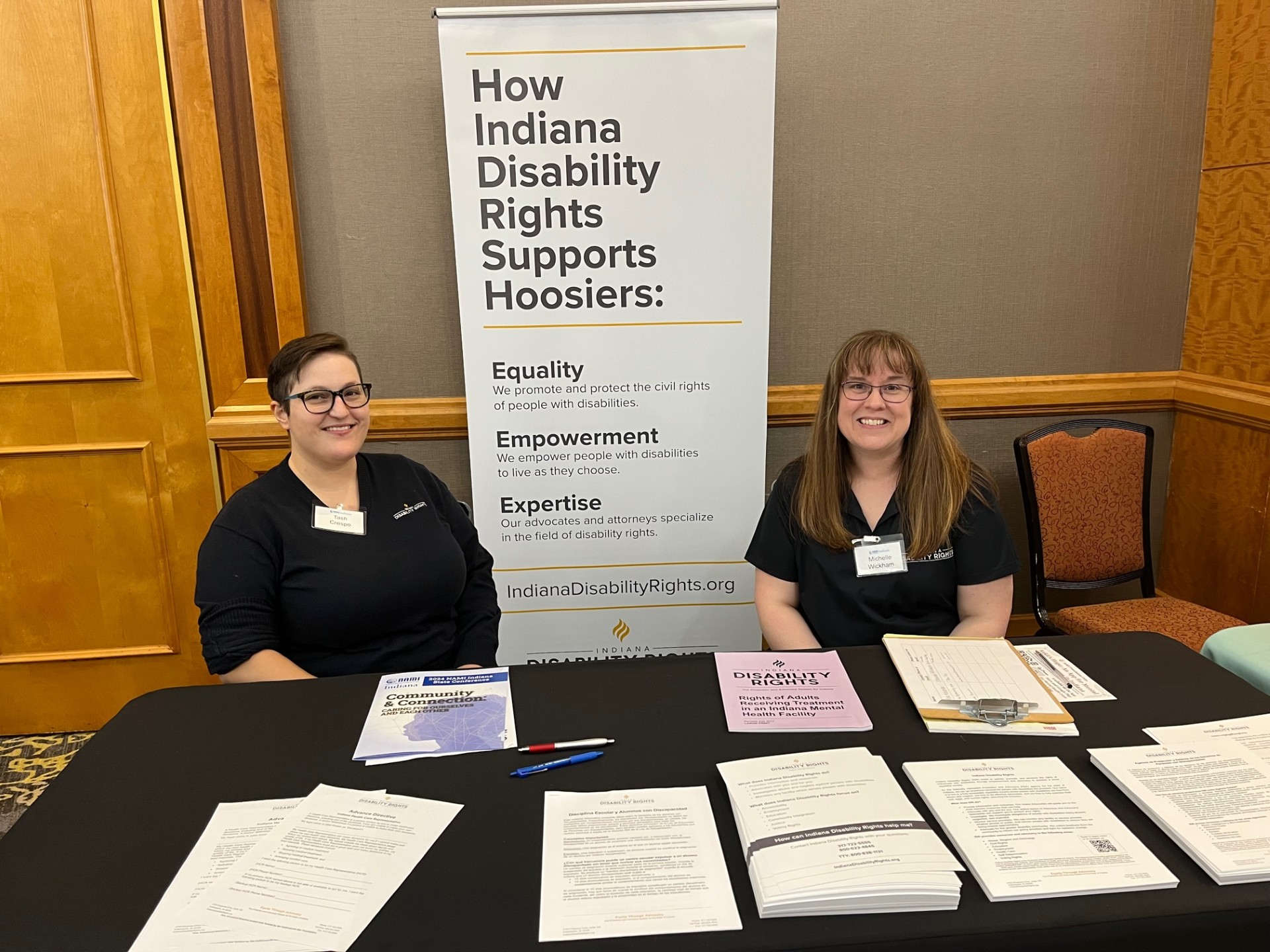 Two people sit at an information table filled with a variety of flyers. A banner stands between them and says, "How Indiana Disability Rights Supports Hoosiers"