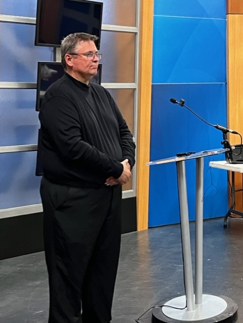 Man in all black clothing stands beside a podium in a television studio setting.