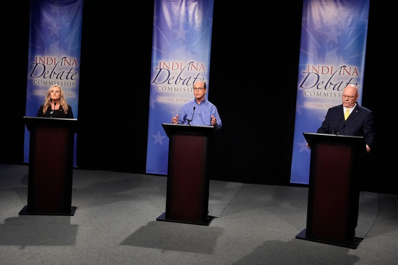 A woman and two men stand behind individual podiums. Blue banners hang behind them.