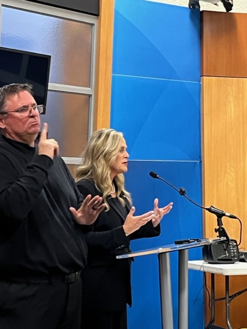 Man in black clothing interprets for a woman in a black suit, who speaks behind a podium.