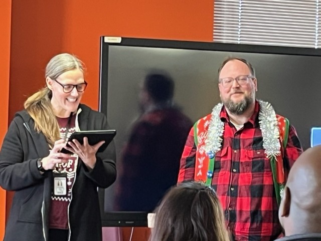 Image of a woman holding a tablet introducing the man next to her to the audience seated in front of them.