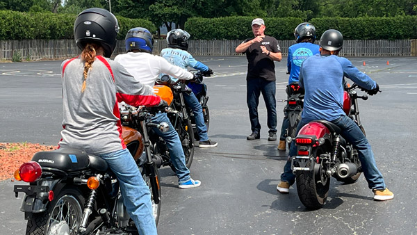 A group of motorcycle students on their bikes, facing the instructor who is giving directions.
